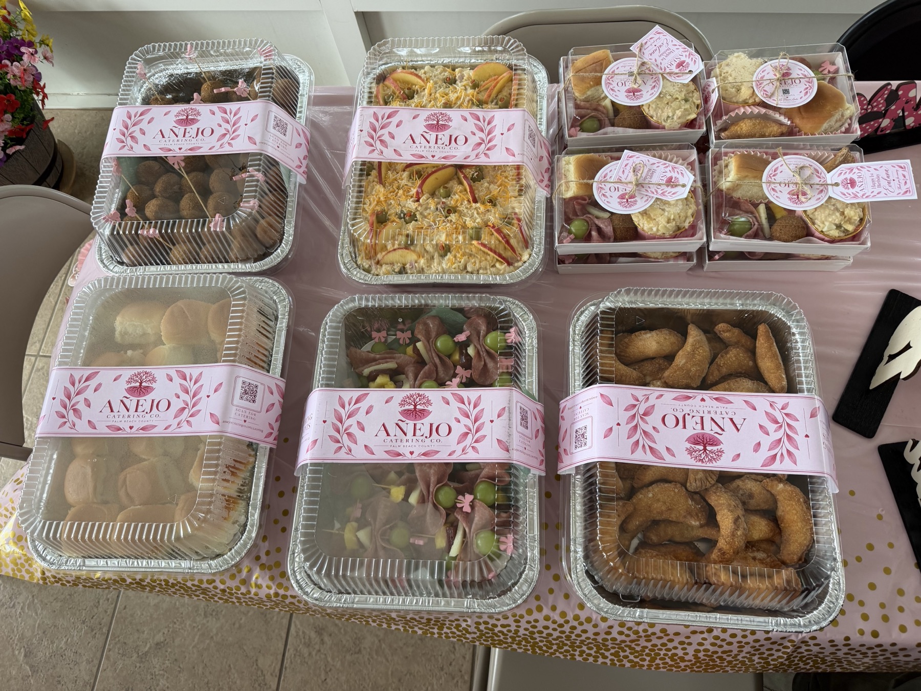 Pink first-birthday catering spread with customized Añejo Cajitas, Hawaiian rolls, savory bites, and party trays